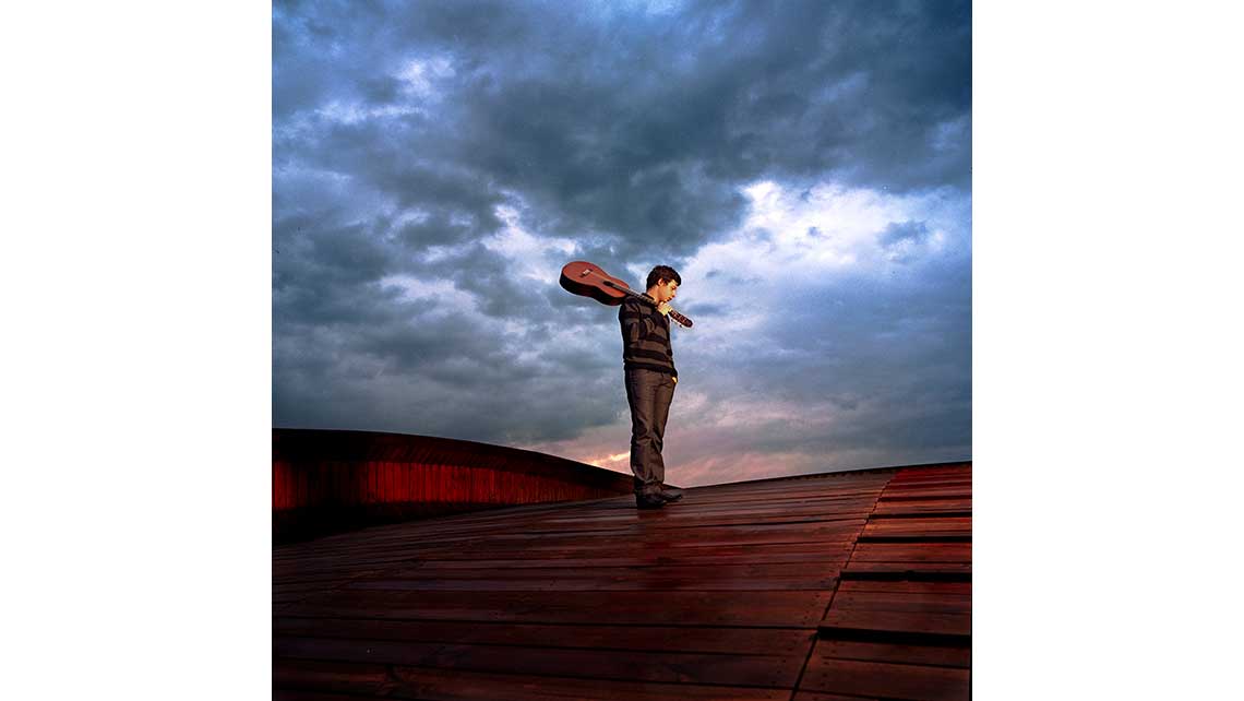 Portrait d'Antoine, élève au Conservatoire de Bordeaux, portant sa guitare sur l'épaule debout sur un toit en forme de voûte et recouvert de bois. © Richard Nourry