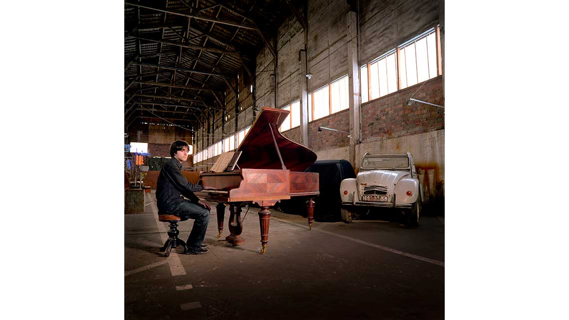 Portrait de Jonathan, pianiste au Conservatoire de Bordeaux, devant son piano placé devant une 2 CV dans un entrepot. © Richard Nourry