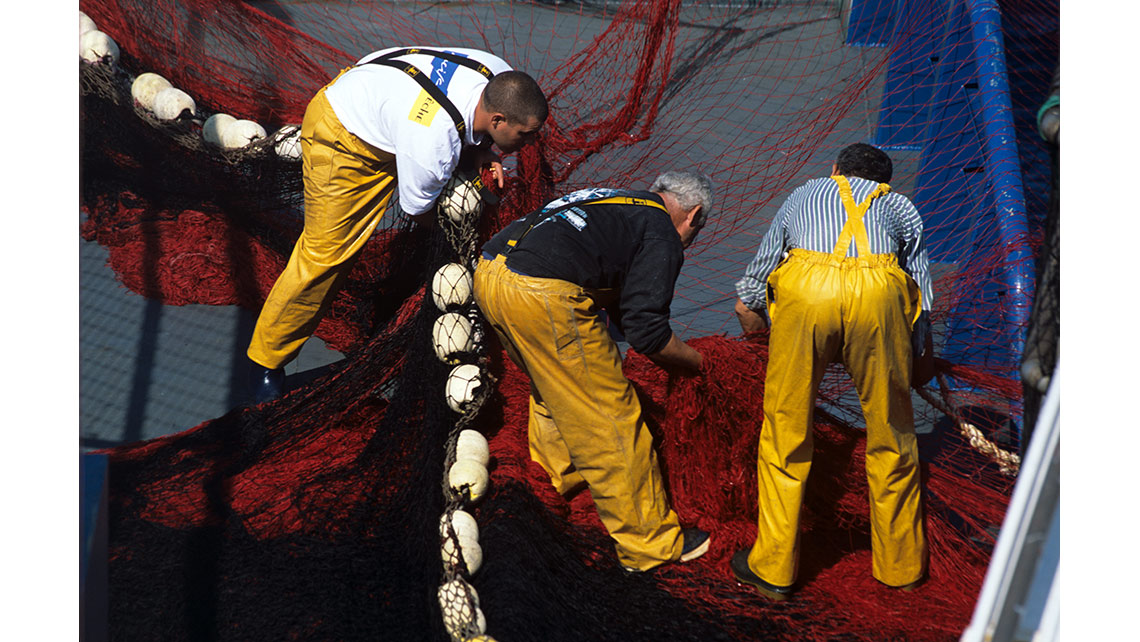 Trois pêcheurs vus de dos sont penchés sur les filets rouges d'un thonier de Sète ; © Richard Nourry