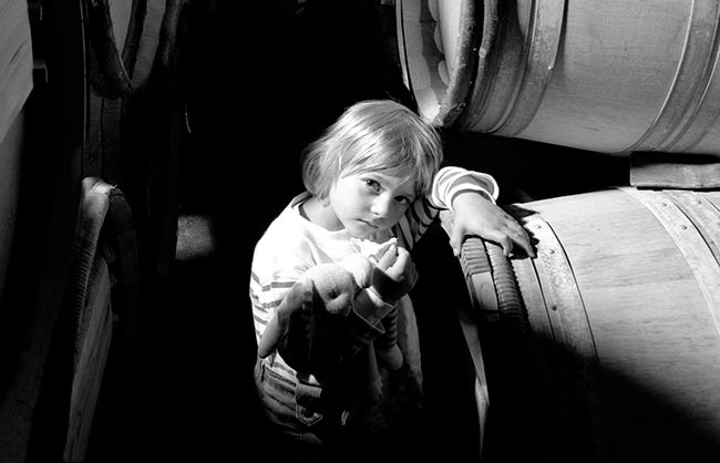 Portrait en noir et blanc d'une petite fille photographiée en plongée entre des fûts de vin dans un chai