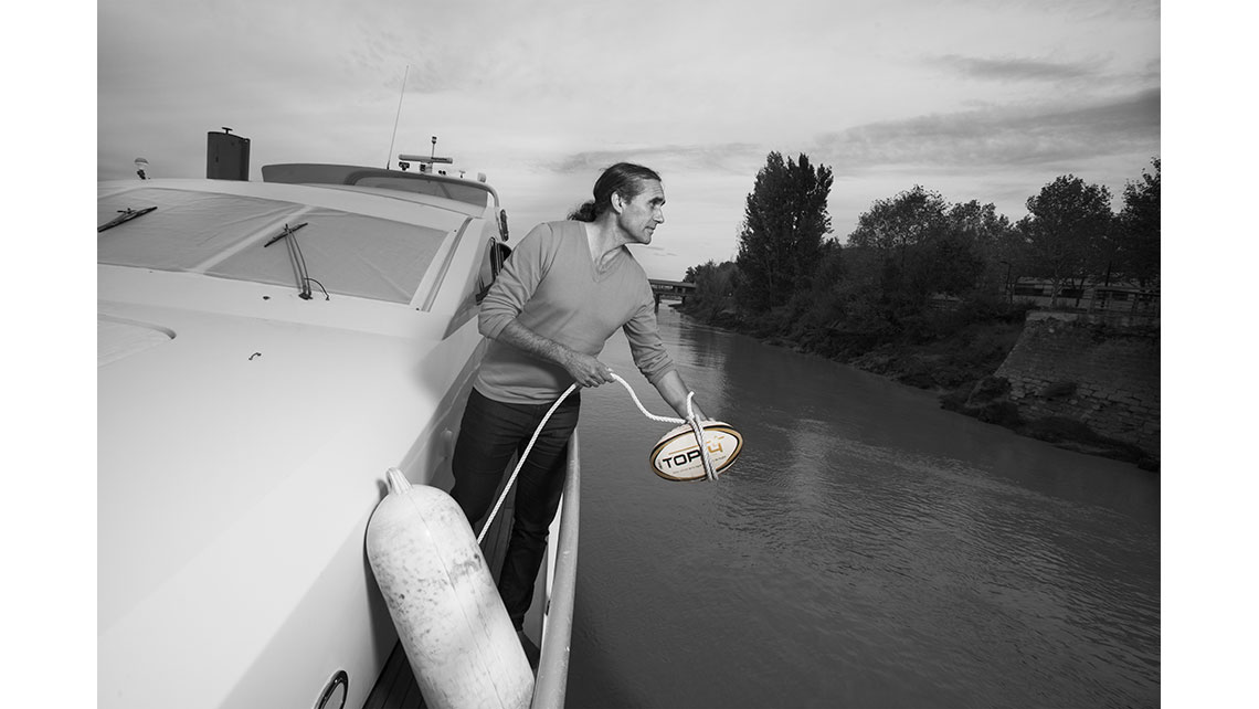 Portrait d'un batelier sur son bateau se préparant à jeter un ballon de rugby remplaçant une bouée d'amarrage, © Richard Nourry